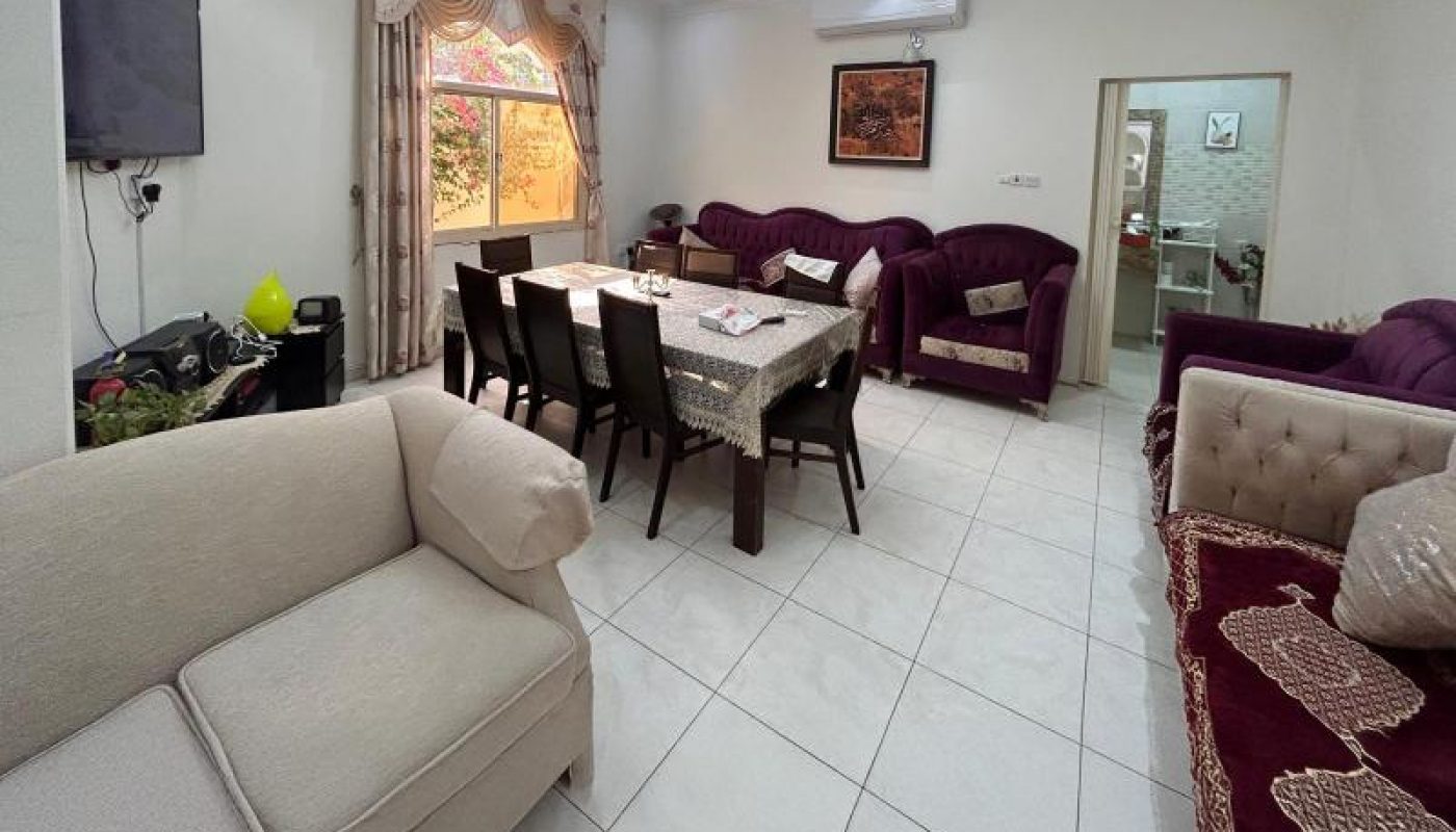 A living room with tiled floor, multiple sofas, a dining table with chairs, a wall-mounted TV, and a doorway leading to another room. A window with curtains lets in natural light.