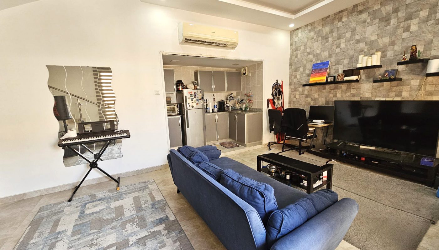 Modern living room with blue sofa, wall-mounted TV, keyboard, rug, and kitchen in the background; patterned accent wall and mirror on the left.