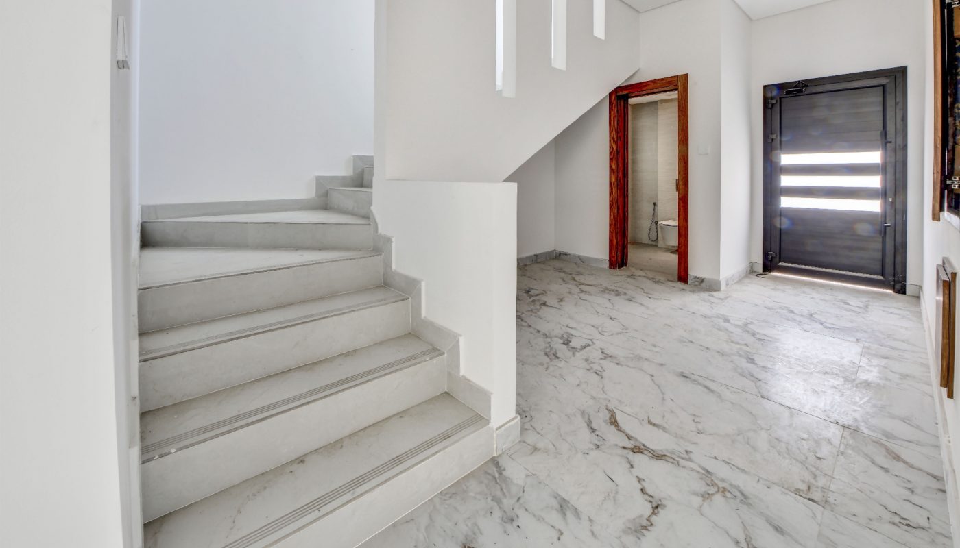 Modern interior with marble floors, white walls, a staircase on the left, a black door on the right, and vertical slit windows allowing natural light.