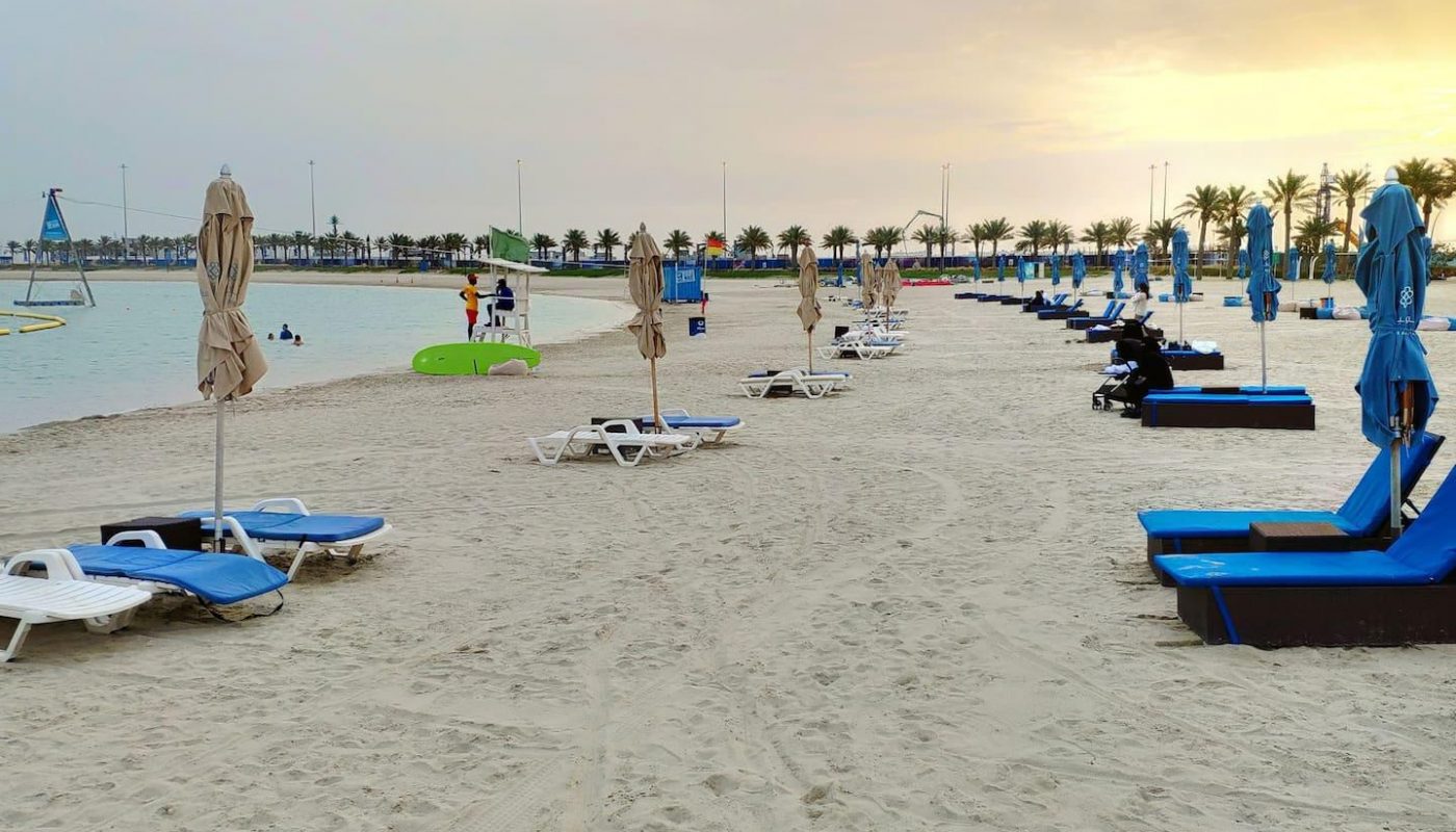 Sandy beach with rows of blue sun loungers and umbrellas, few people in the distance, palm trees, and calm sea under a cloudy sky.