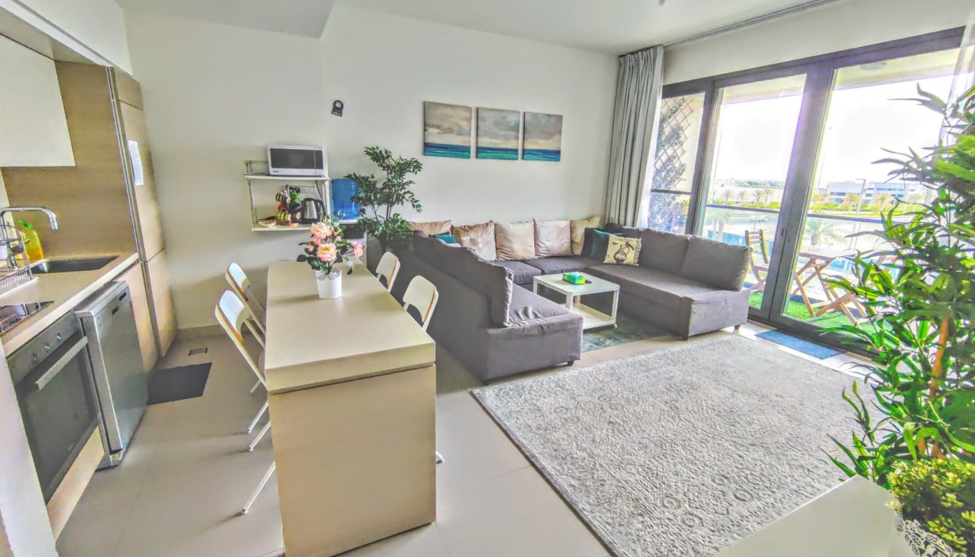 Modern open-plan living room and kitchen with a corner sofa, dining table, indoor plants, and large windows leading to a balcony with outdoor seating.