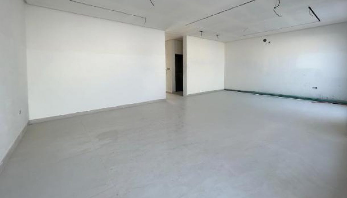 An empty, unfinished room with white walls, a grey floor, exposed wiring on the ceiling, and a single open doorway in the back wall.