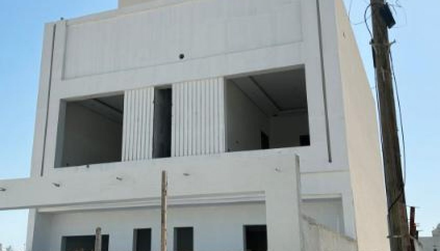A modern, white, two-story building under construction, partially obscured by a tall wooden fence, with a utility pole nearby and a clear blue sky above.