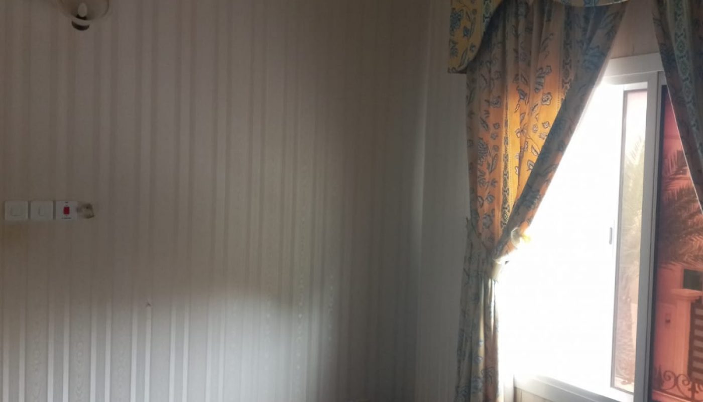 An empty room with blue carpet, striped wallpaper, a window with floral curtains, a plastic storage box, and a wall light fixture.