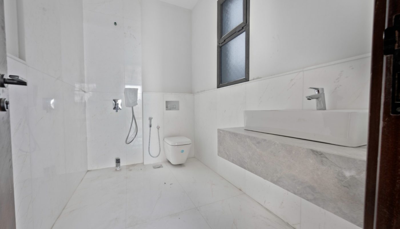 Modern bathroom with white marble walls and floor, wall-mounted toilet, bidet shower, rectangular sink, and frosted window.