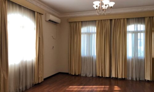 A 5-bedroom villa located in the Janbyah area, featuring a room with wooden flooring, three windows adorned with cream curtains, an air conditioner on the wall, and a ceiling light fixture. This semi-furnished property offers comfort and elegance.