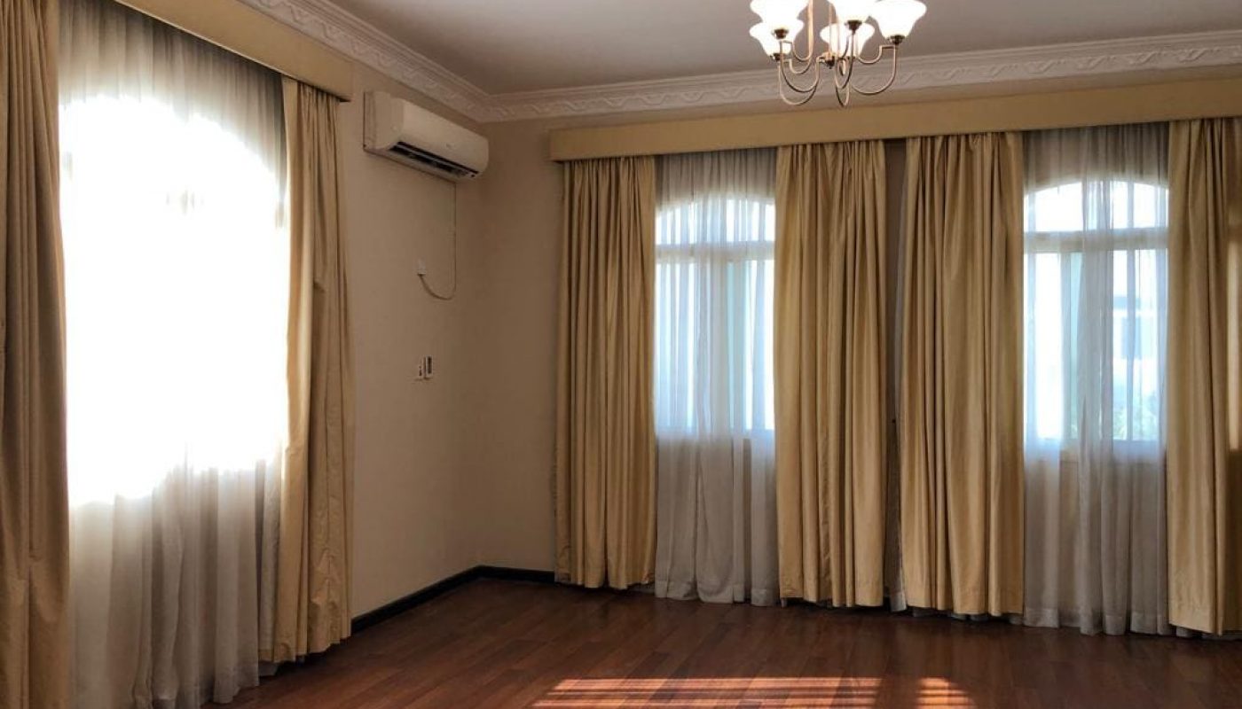 A 5-bedroom villa located in the Janbyah area, featuring a room with wooden flooring, three windows adorned with cream curtains, an air conditioner on the wall, and a ceiling light fixture. This semi-furnished property offers comfort and elegance.