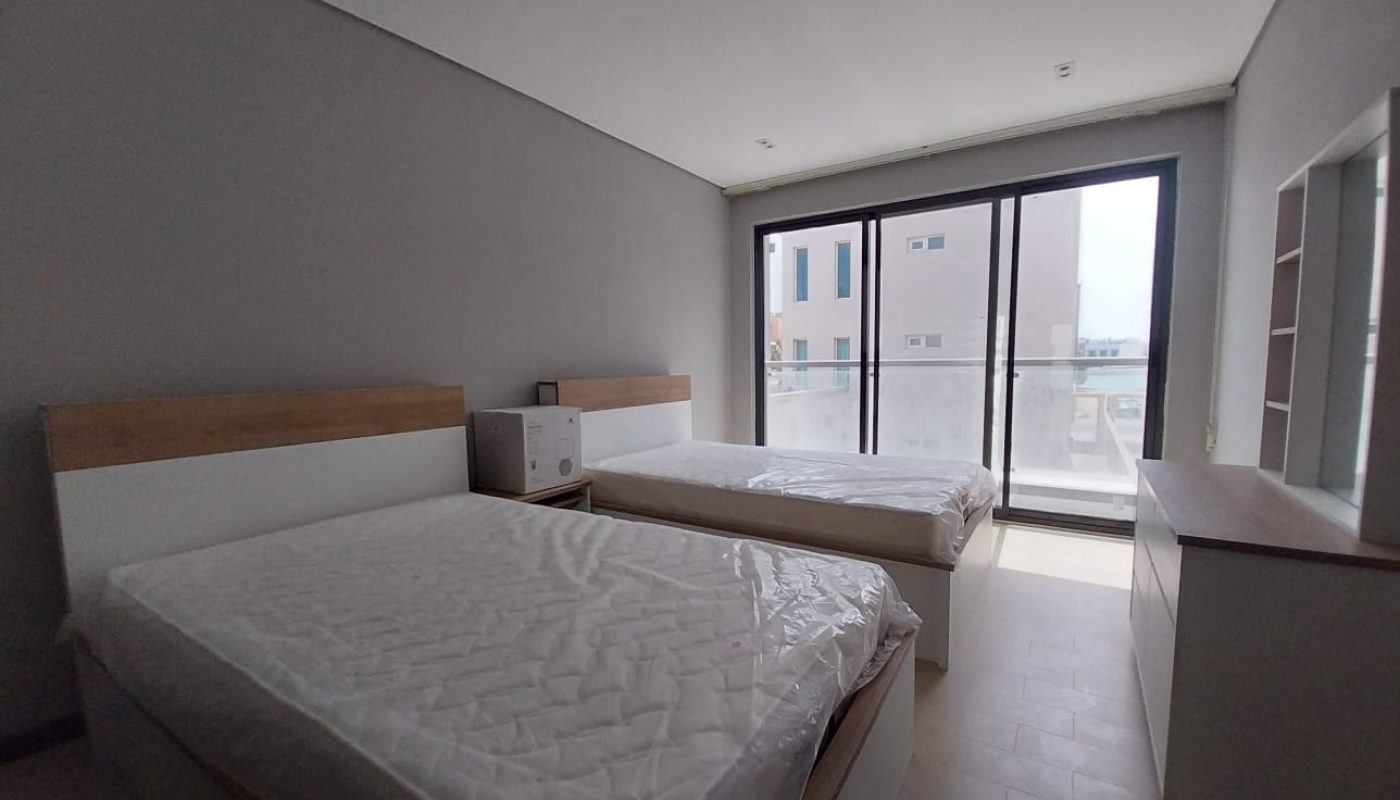 A bright bedroom with two single beds, each covered with plastic-wrapped mattresses. A window and glass door provide natural light, overlooking a building. A dresser is on the right.