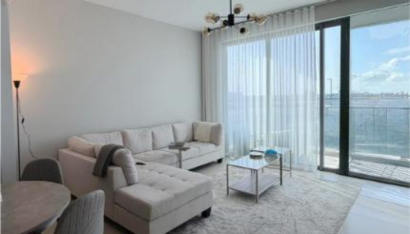 Modern living room with light-colored furniture, a sectional sofa, coffee table, and rug; large windows with sheer curtains overlook a balcony and water view.
