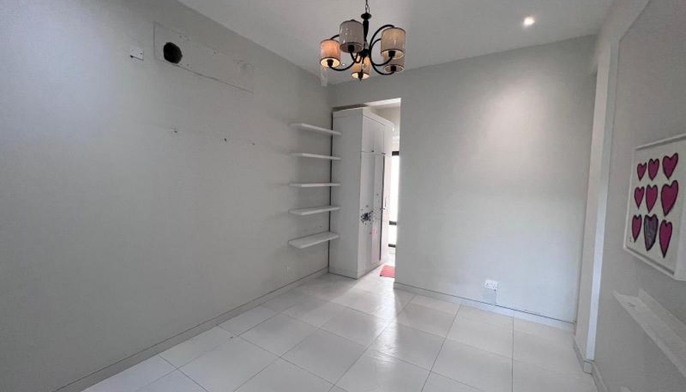 A small, empty room with white tiled floor, light gray walls, wall-mounted shelves, a chandelier, and a door leading to another space. A heart-themed artwork hangs on the right wall.