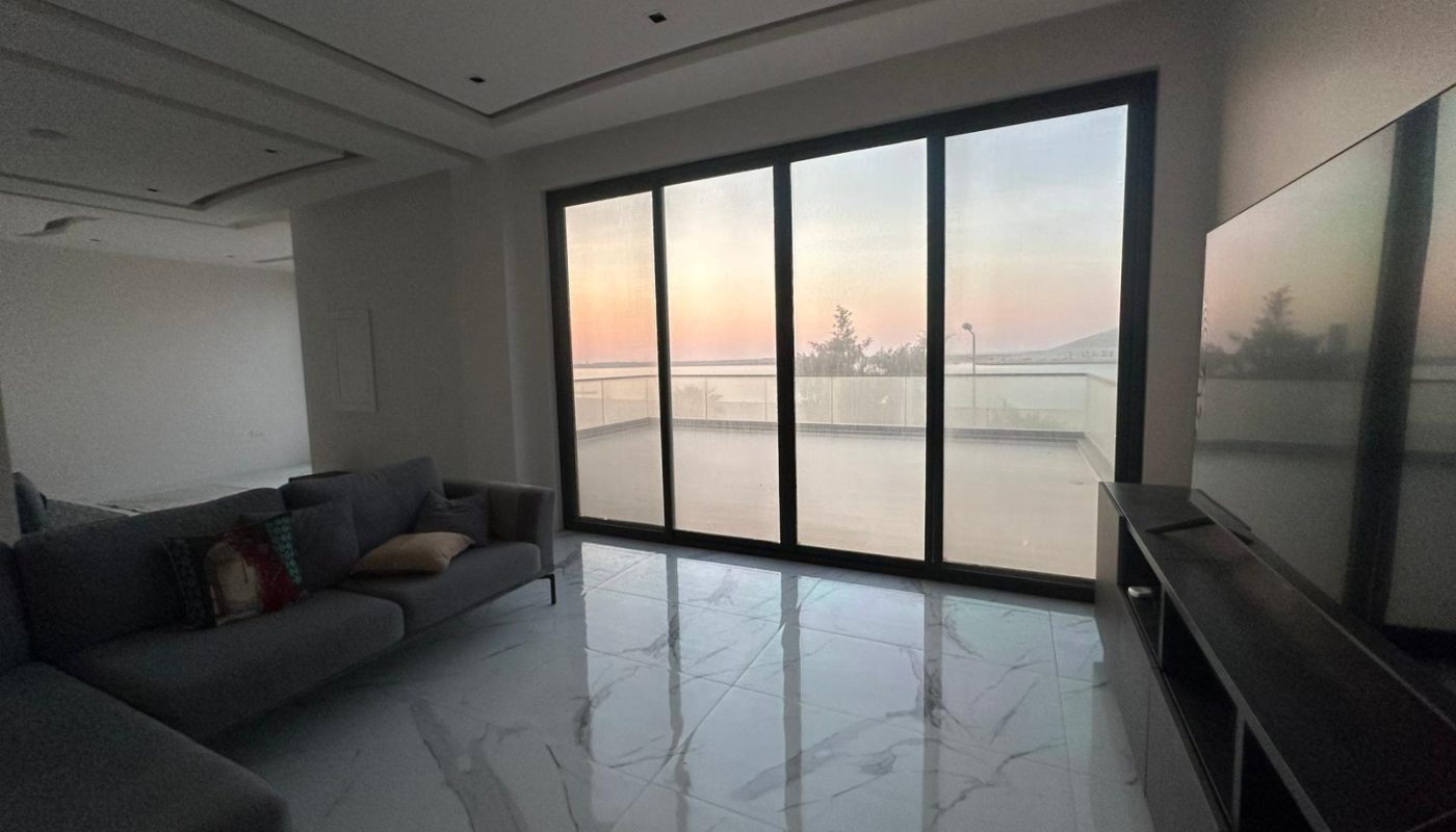 Modern living room with a large gray sofa, marble tile floor, and a flat-screen TV, featuring floor-to-ceiling windows overlooking a terrace and a sunset view.