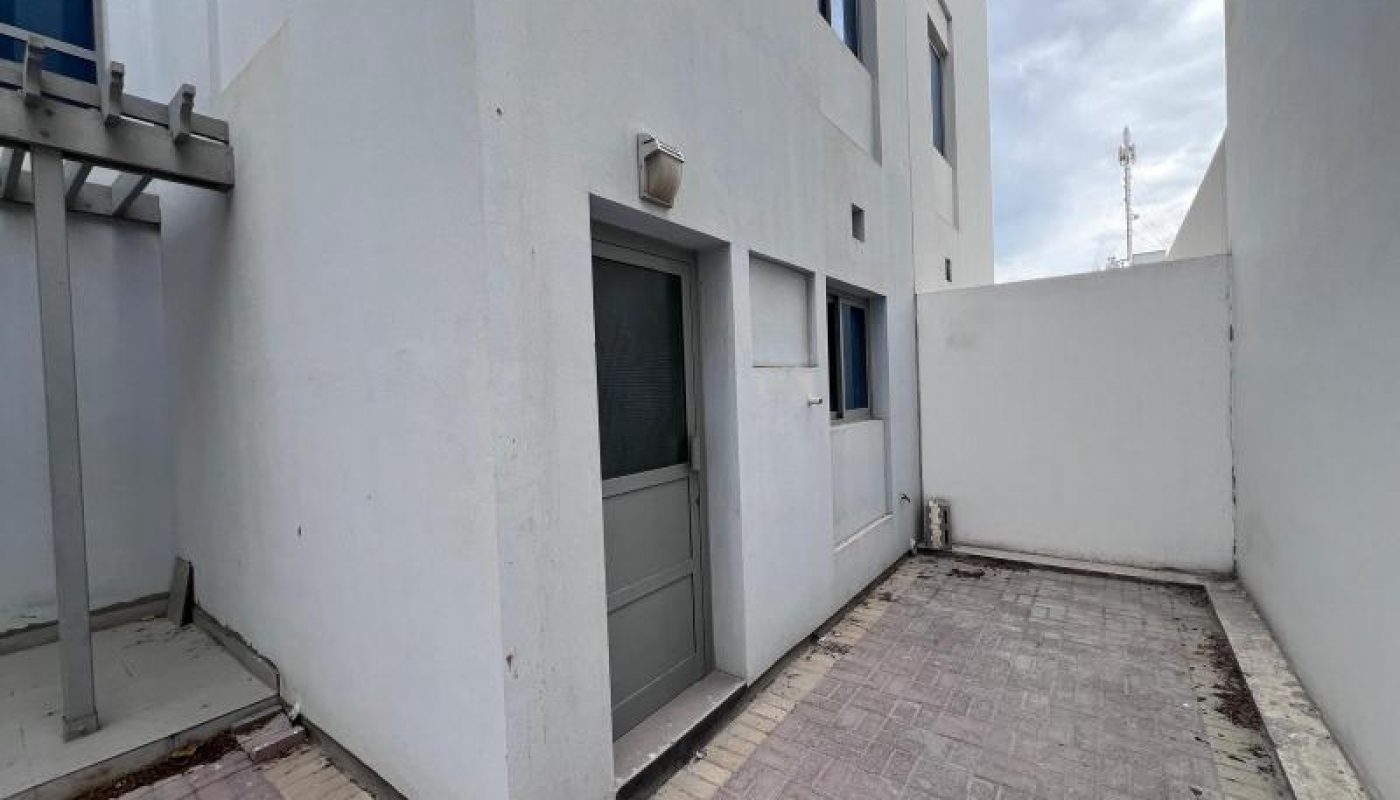 A small concrete courtyard with light gray paving stones, adjacent to a white building with a gray door and two windows; overcast sky above.
