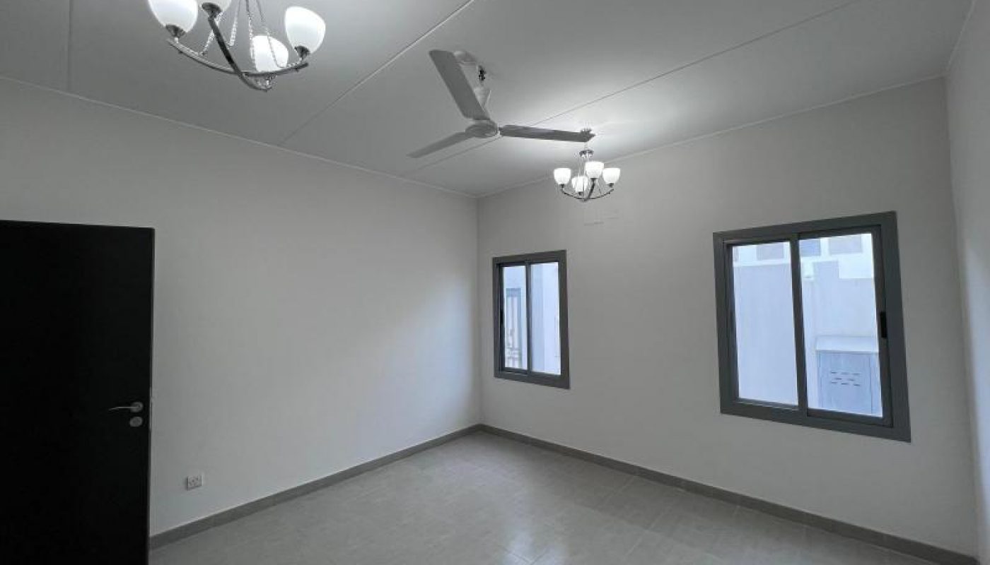 Empty room with white walls, tiled floor, two windows, a ceiling fan, and two modern light fixtures. The door is open and natural light enters through the windows.