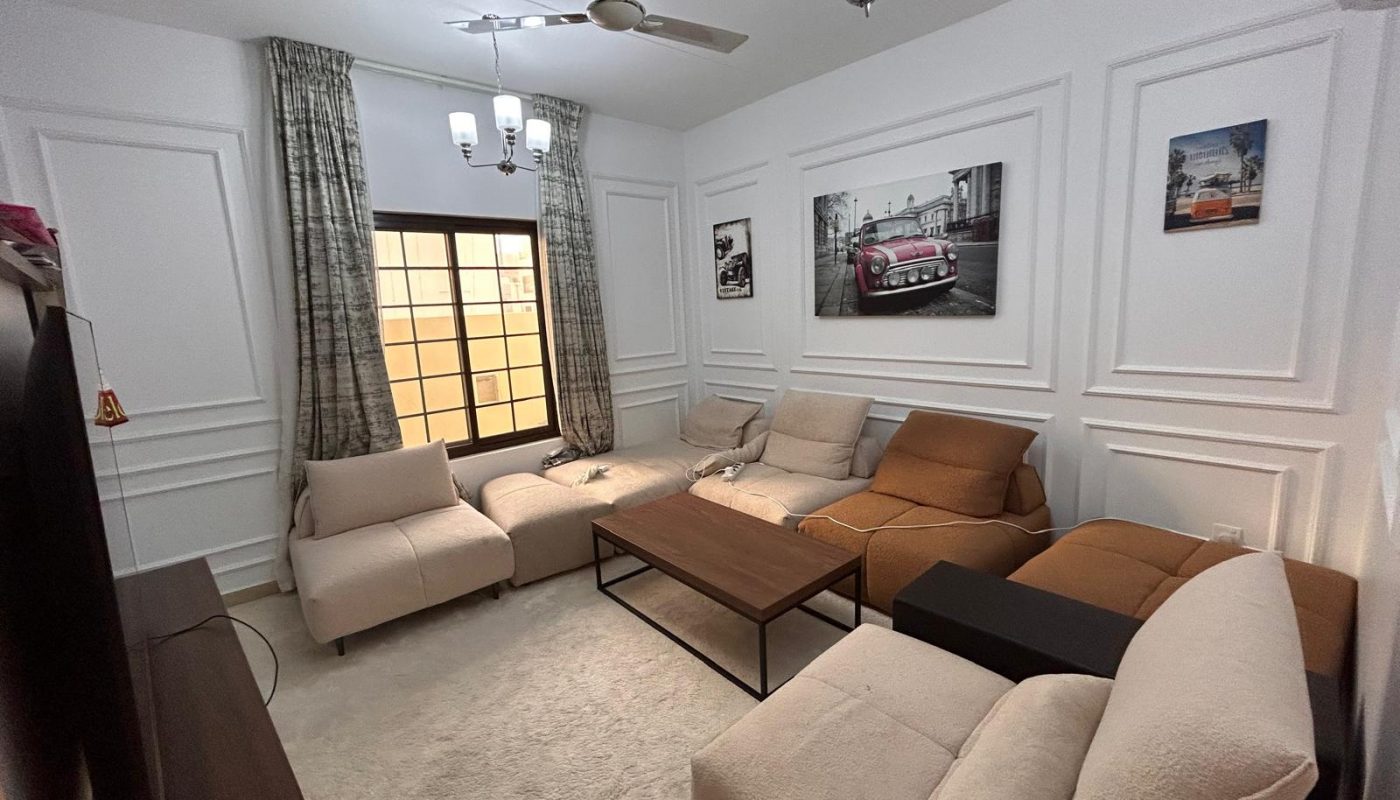 A modern living room with beige and brown sofas, a wooden coffee table, wall art of cars, ceiling fan, light fixtures, and a window with patterned curtains.