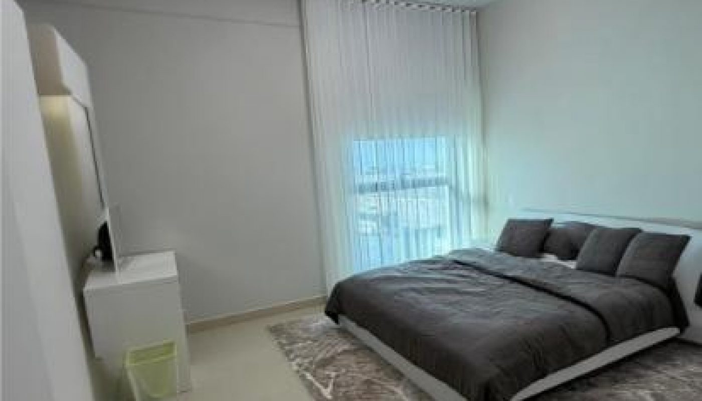 Minimalist bedroom with a large window, gray bedding, a modern chandelier, a white desk, wall-mounted TV, and a neutral-toned rug on a light floor.