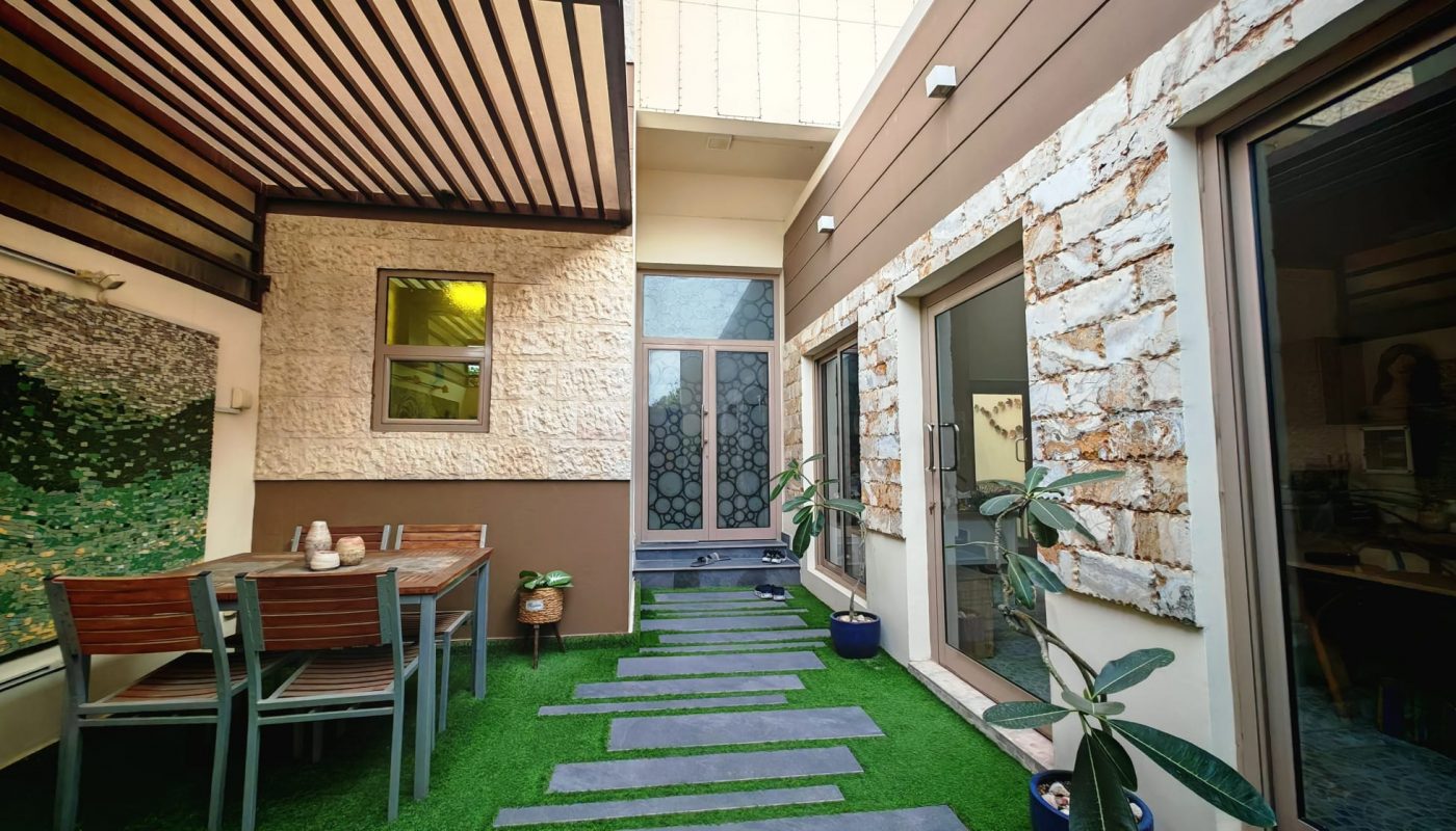 A modern patio with a table and chairs, potted plants, stone and wood walls, and stepping stones on artificial grass leading to a glass door.