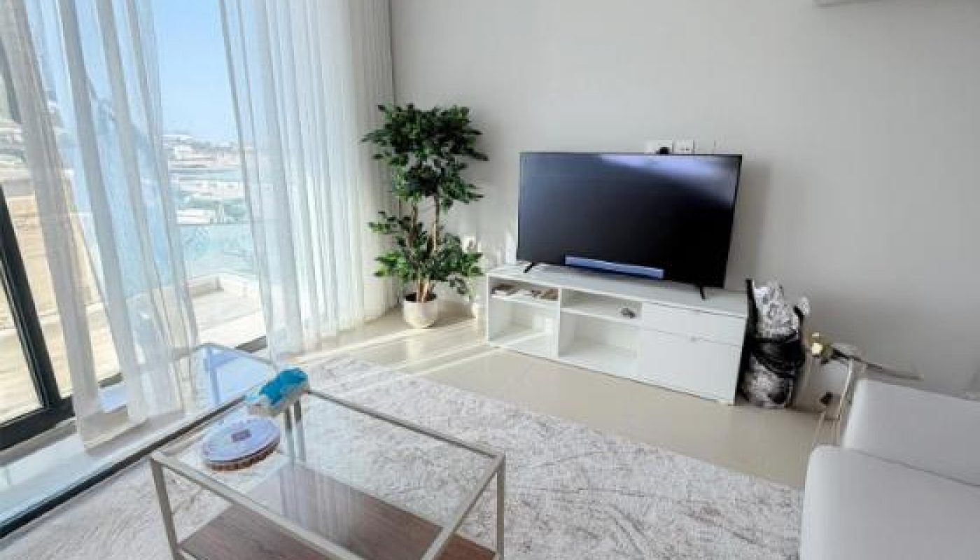 A modern living room with a glass coffee table, white rug, TV on a white stand, potted plant, large window with sheer curtains, and an air conditioner on the wall.