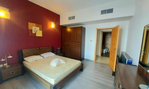 A well-lit flat in Amwaj featuring a large bed with white bedding against a deep red wall, wooden flooring, an open door, and minimalistic decor.