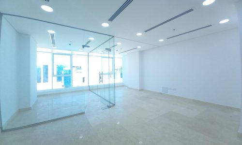 An empty office with glass walls and a glass door in a bright building in the heart of Seef.