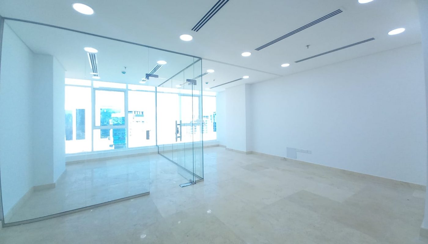 An empty office with glass walls and a glass door in a bright building in the heart of Seef.