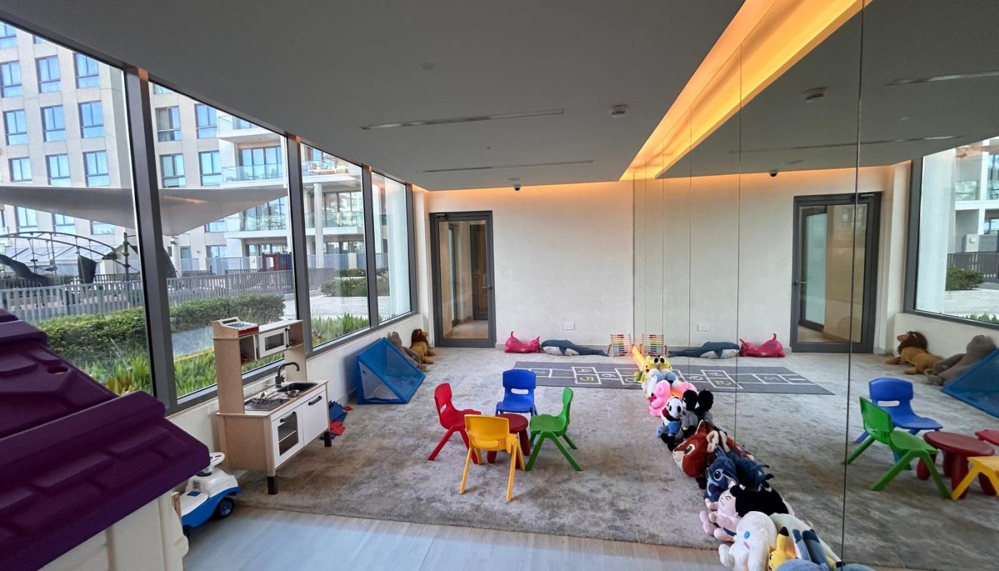 Indoor children's playroom with small colorful chairs, stuffed animals lined up against a mirrored wall, playhouses, and large windows overlooking an outdoor area.