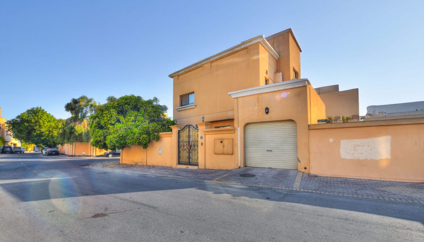 A two-story tan house with a garage and gated entrance, located on a sunny street corner with trees nearby and a clear blue sky overhead.