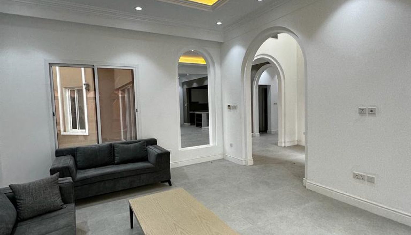 Modern living room with gray sofas, a light wood coffee table, arched doorways, recessed ceiling lighting, and white walls with tile flooring.