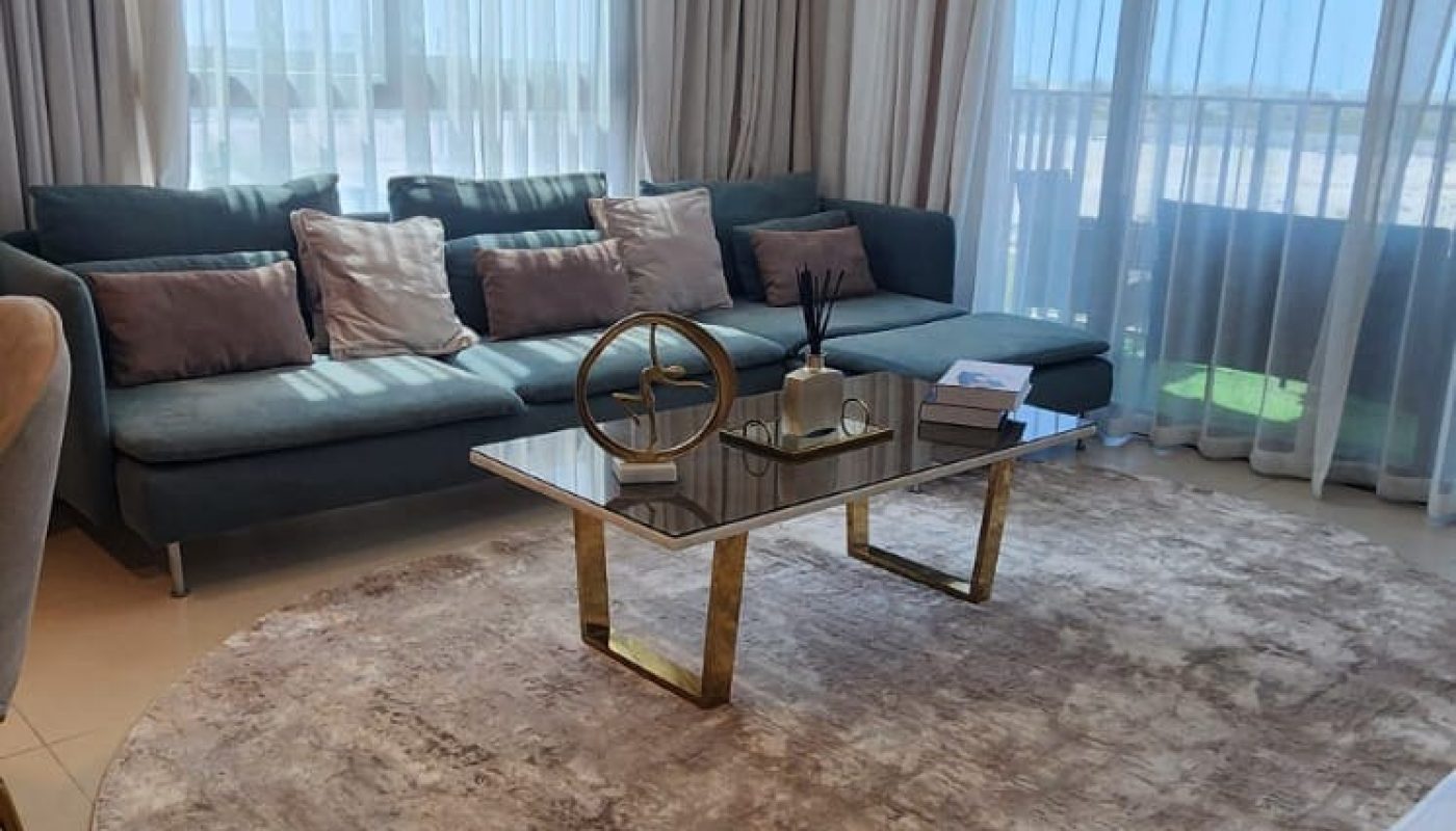 A modern living room with large windows, sheer curtains, a chandelier, a round rug, a coffee table, and a gray sofa with cushions. A plant is in the foreground.