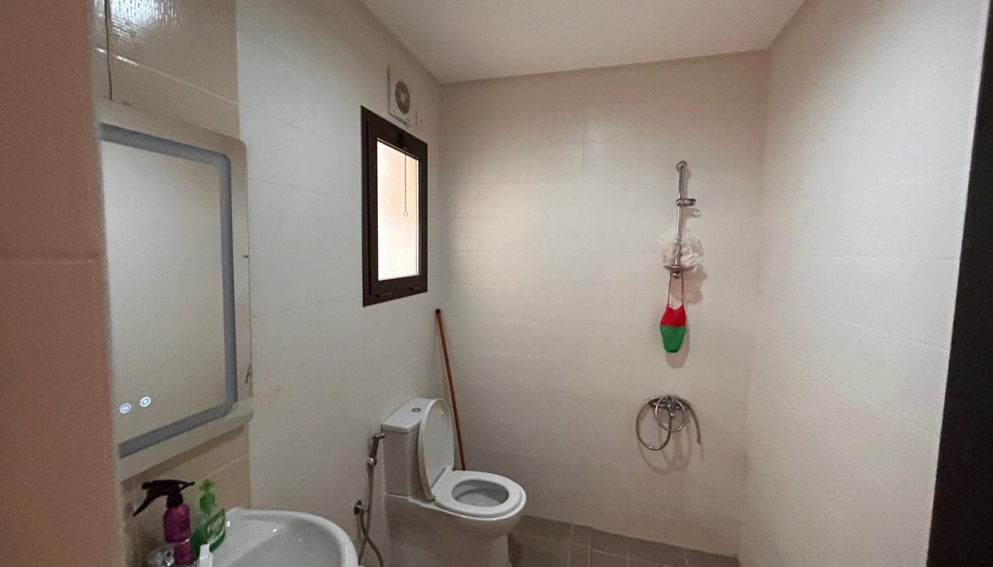 A bathroom with a sink, mirror, toilet, window, wall-mounted shower, cleaning supplies, and a mop against the wall.