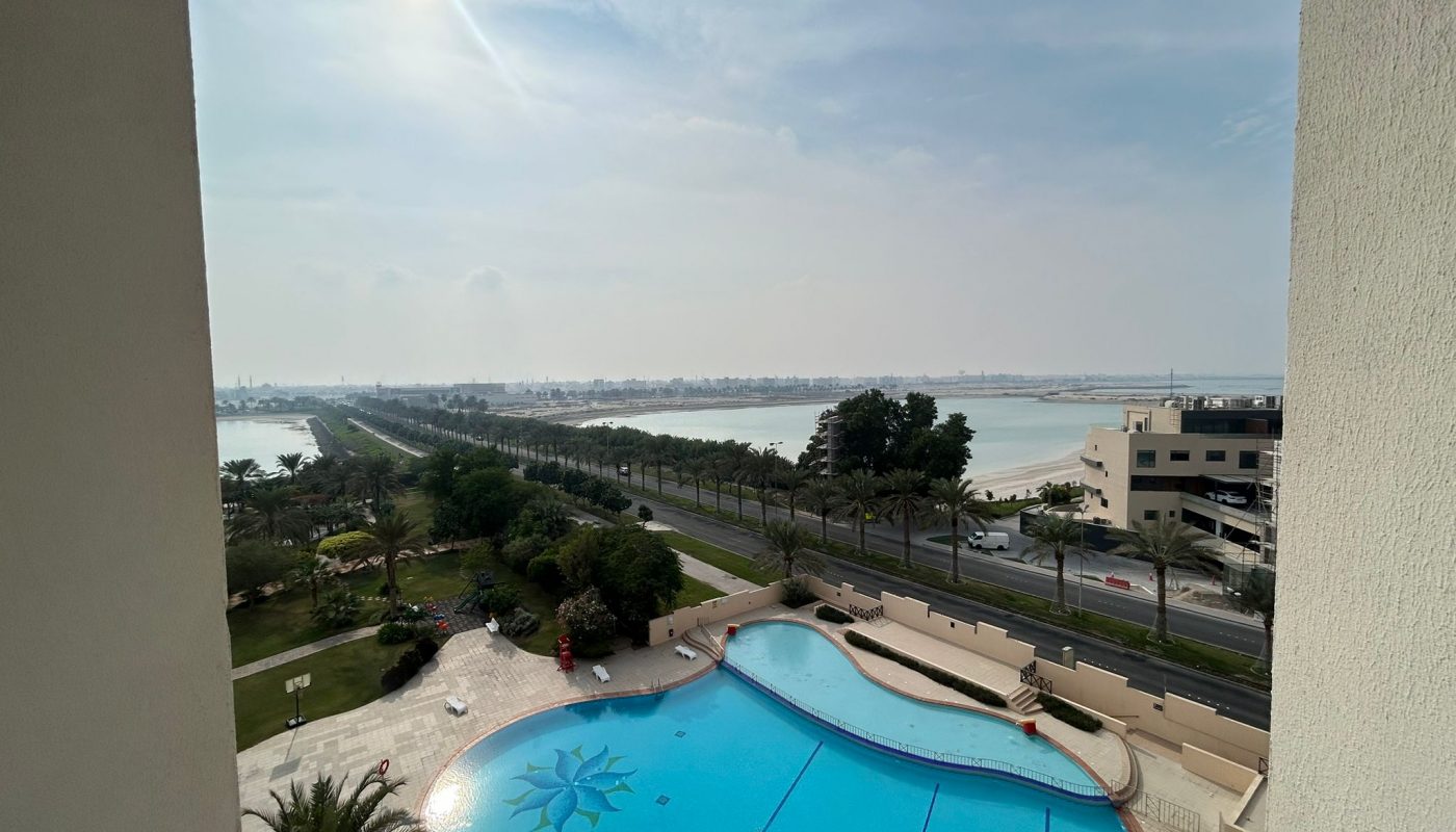 View from a balcony showing a swimming pool with a sun motif, palm trees, a road, and a body of water in the distance under a partly cloudy sky.