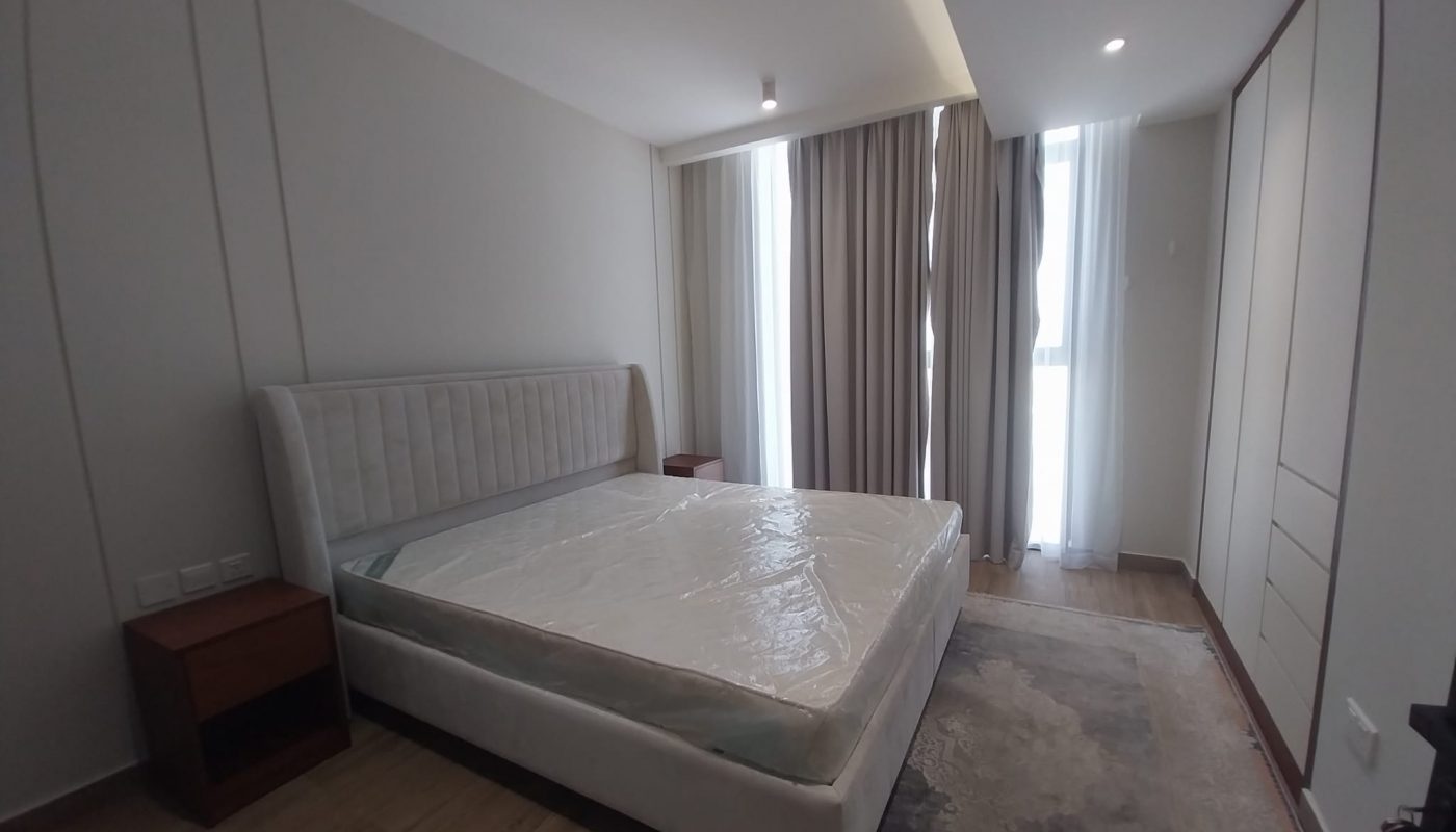 A modern bedroom with a large bed covered in plastic, two nightstands, floor-to-ceiling curtains, and built-in wardrobes along one wall.