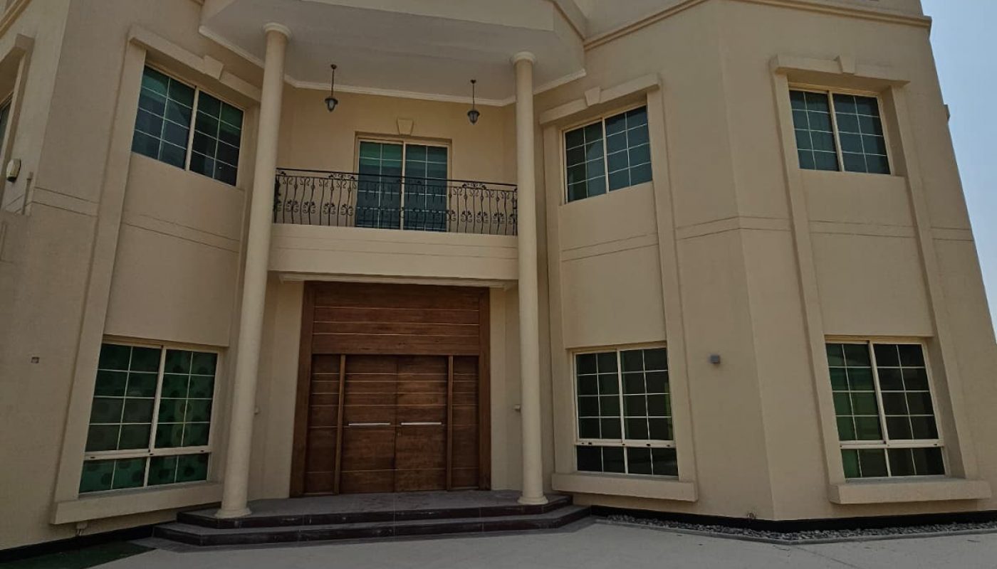 A large, beige, two-story building with green-tinted windows, a wooden double door entrance, and a balcony above the main entrance under a clear sky.