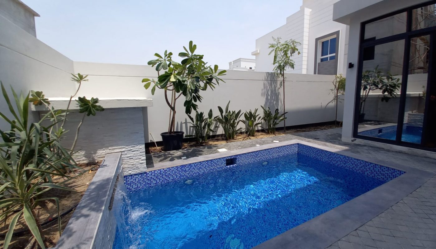 A small rectangular pool with blue tiles is surrounded by plants and trees in a modern walled backyard next to a glass door.