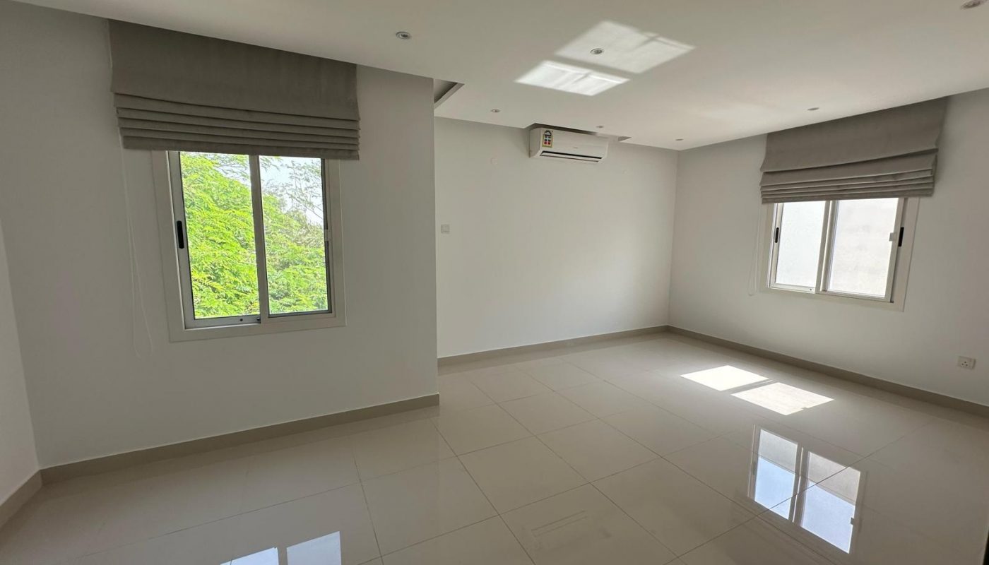 Empty, unfurnished room with white walls, tiled floor, two windows with gray blinds, and a wall-mounted air conditioner. Sunlight is coming through the windows.