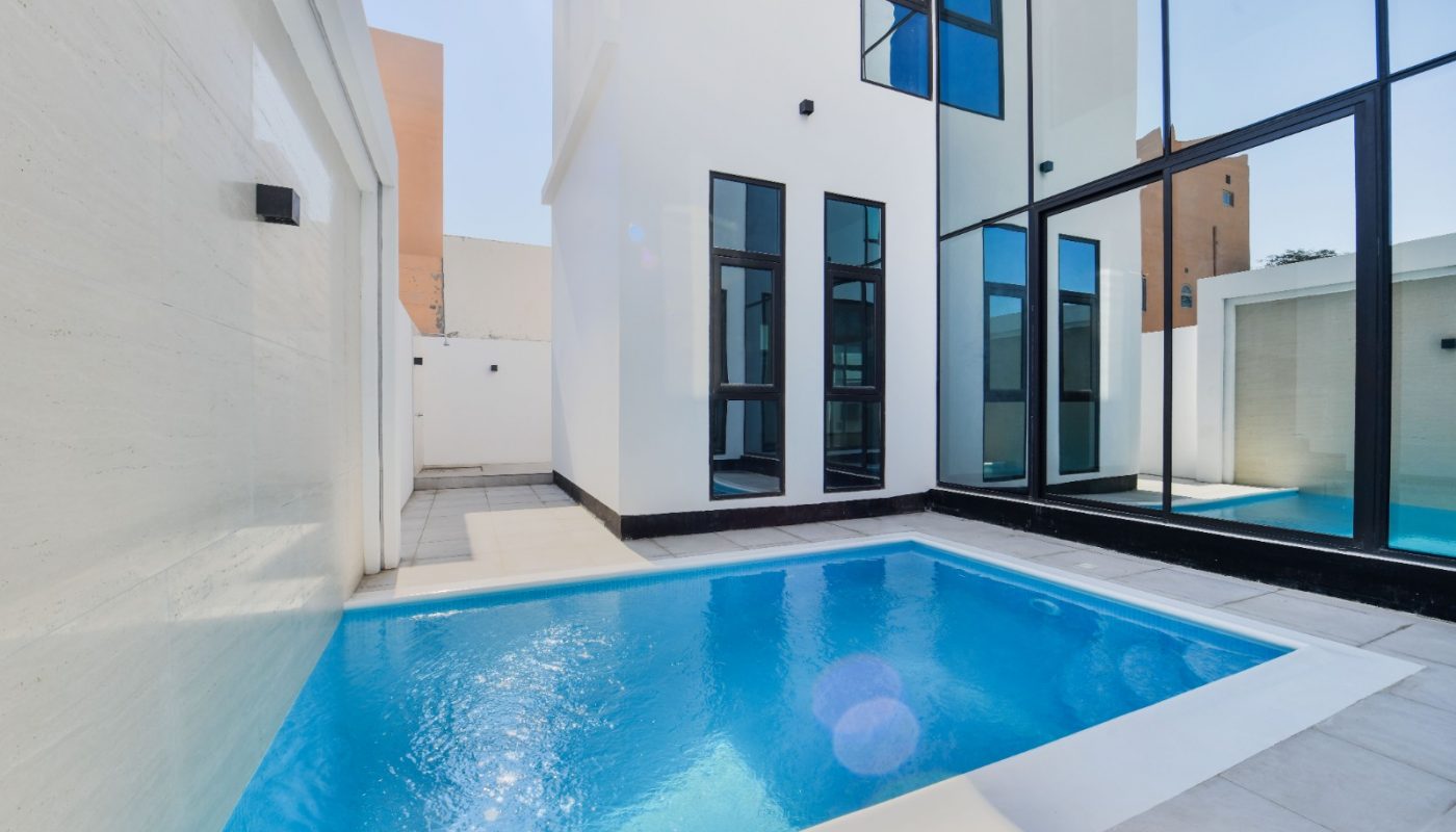 Rectangular swimming pool beside a modern white building with large floor-to-ceiling windows and a tiled patio area.