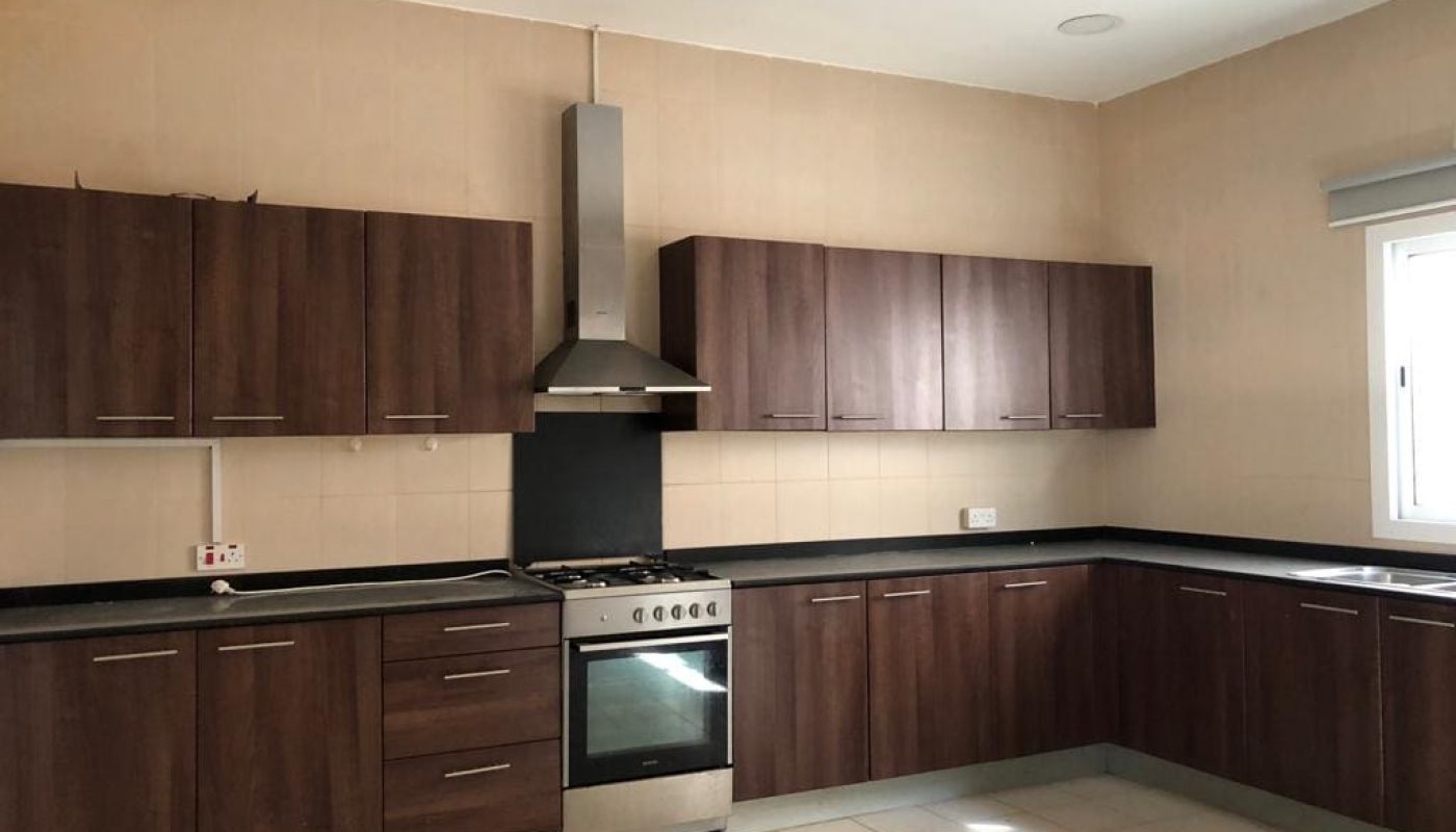 A modern kitchen with wooden cabinets, a stainless steel stove and oven, a range hood, and a window providing natural light. The countertops are black, and the floor is tiled in a light color.