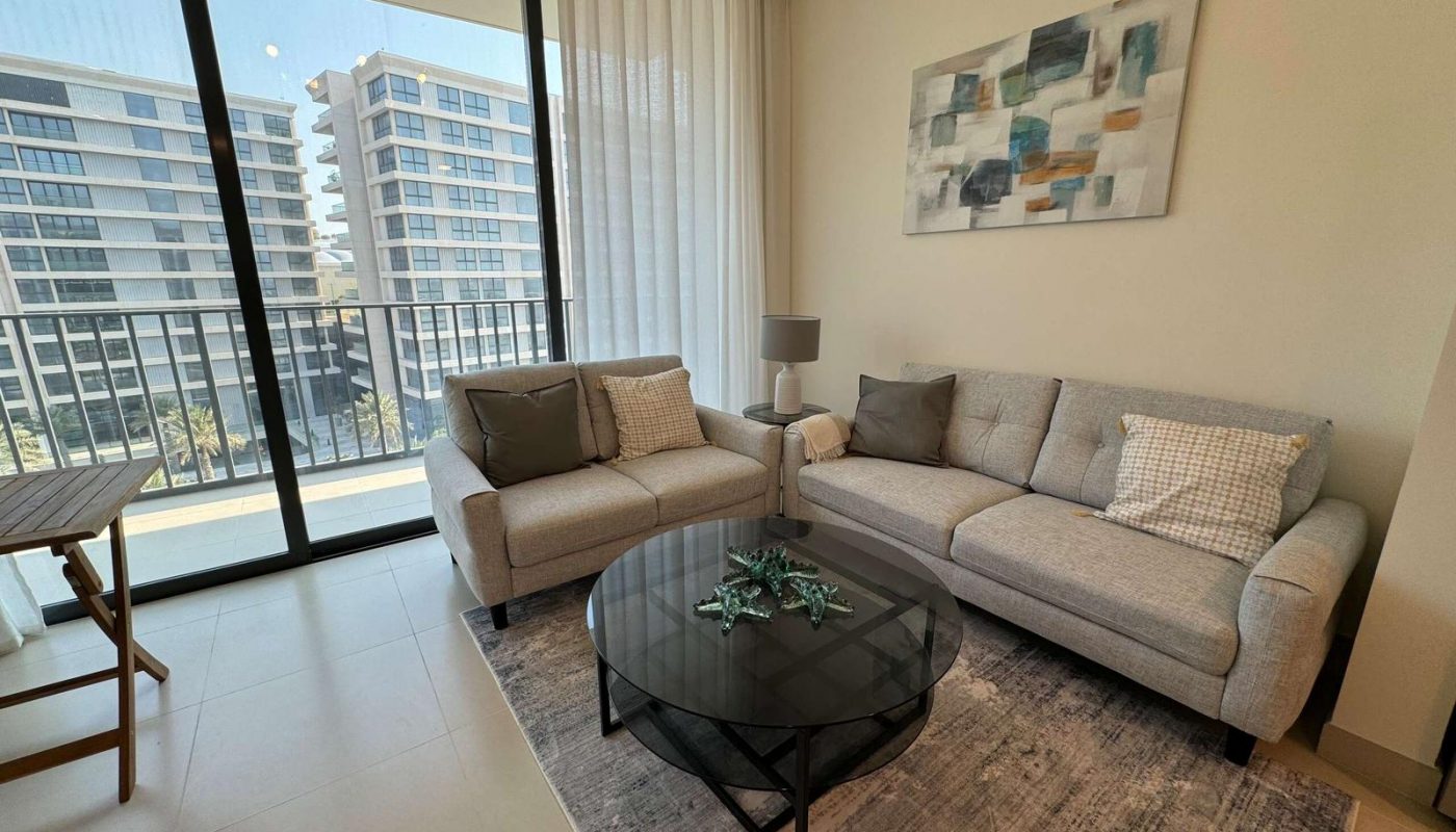 Modern living room with two gray sofas, a glass coffee table, abstract wall art, and a balcony view of apartment buildings through floor-to-ceiling windows with sheer curtains.