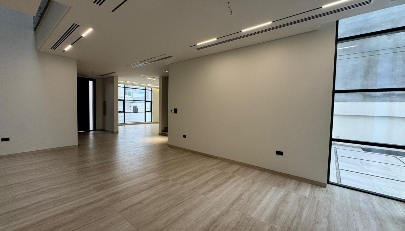 Spacious, modern empty room with light wood floors, white walls, recessed lighting, and large windows letting in natural light.