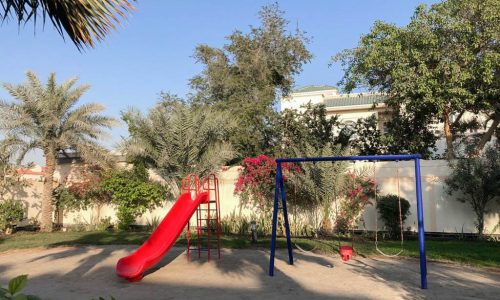 A luxury playground with a red slide, palm trees, garden, and private pool.