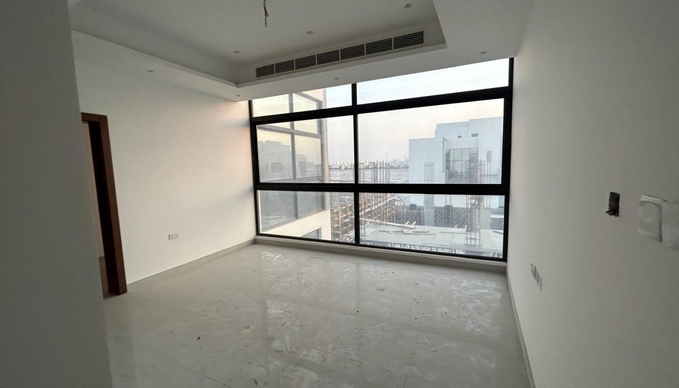 Empty, unfurnished room with large floor-to-ceiling windows, white walls, and a tiled floor overlooking buildings outside. Ceiling lights and duct vents are visible.