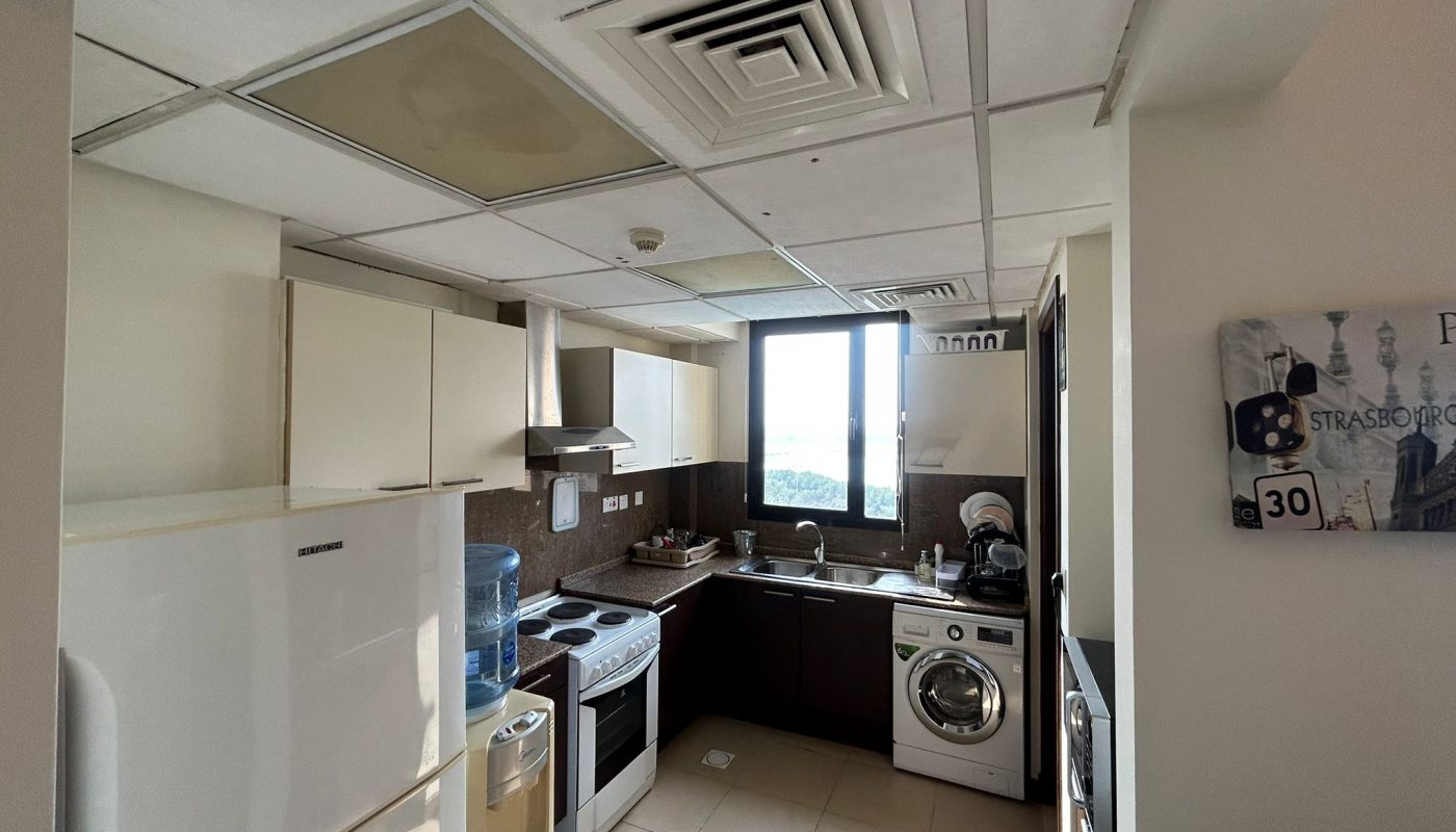 A compact kitchen with white cabinets, stove, washing machine, refrigerator, and water dispenser; sunlight comes through a window above the sink.