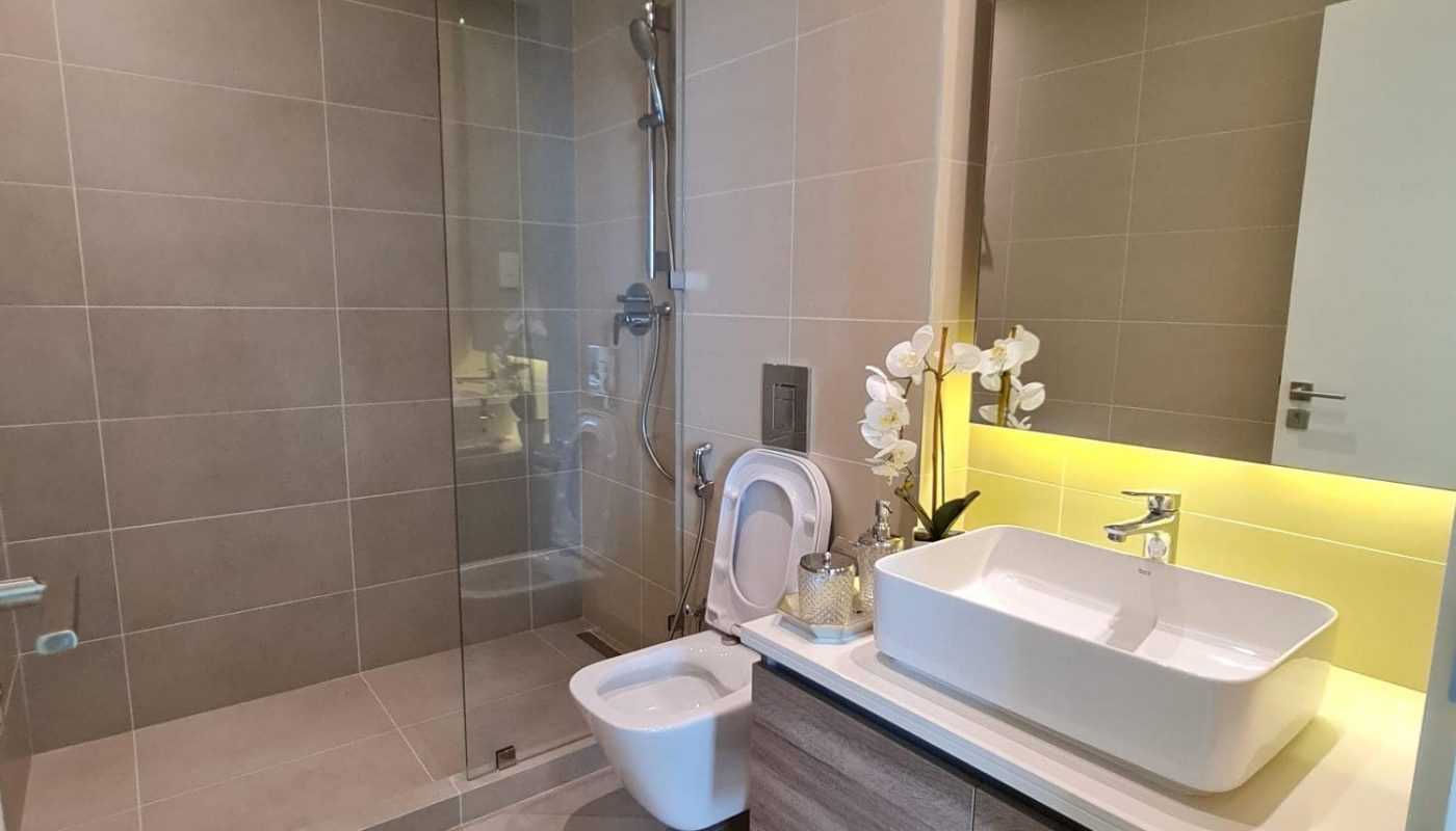 Modern bathroom with a glass shower, white toilet, rectangular sink on a wooden vanity, and a backlit mirror. Neutral tiles and an orchid plant decorate the space.