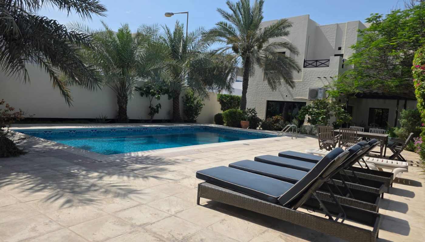 Four black lounge chairs are lined up beside a rectangular pool, surrounded by palm trees and a modern white building under a clear sky.