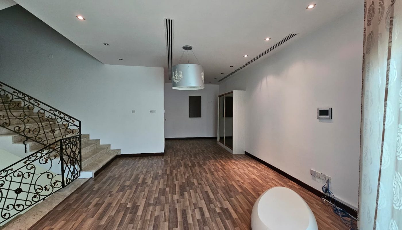 A modern, empty living room with wood flooring, a staircase with ornate railings, a white round ottoman, and large windows with curtains. Ceiling lights and a hanging fixture are visible.