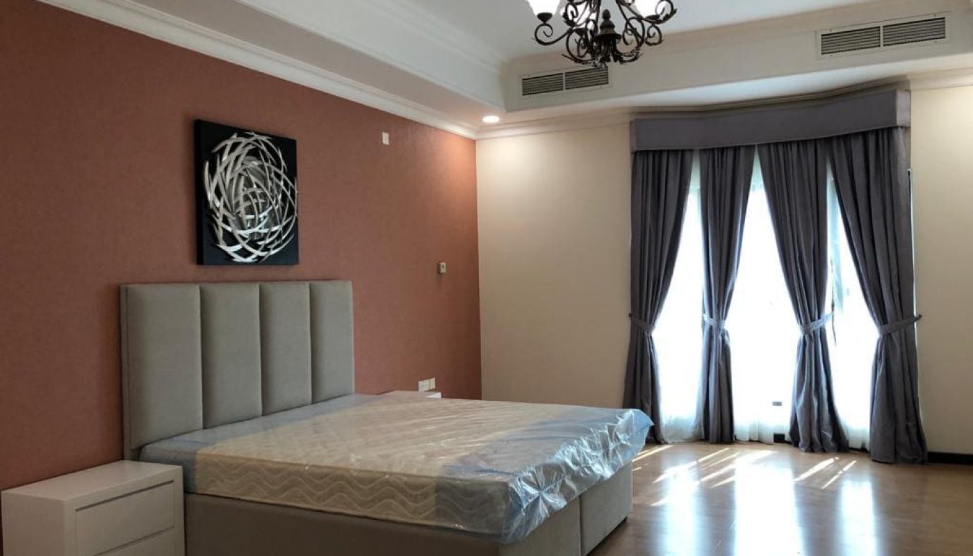A modern, fully-furnished bedroom with a large bed, light gray headboard, terracotta walls, and a geometric wall art piece above the bed. White curtains frame a window.
