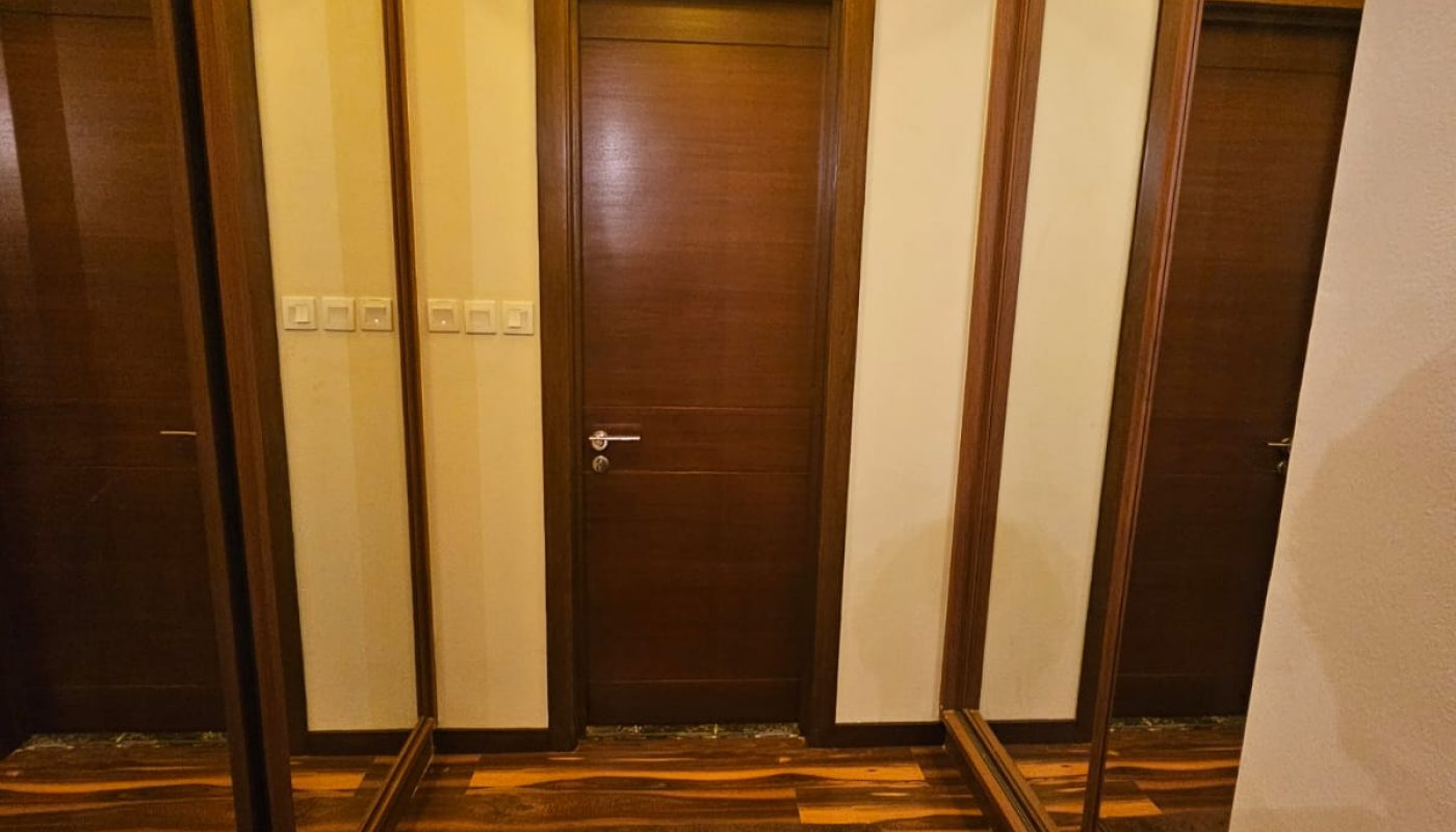 A hallway with a closed wooden door, brown floor mats, and floor-to-ceiling mirrors on both sides reflecting the door and walls. Warm ceiling light illuminates the space.