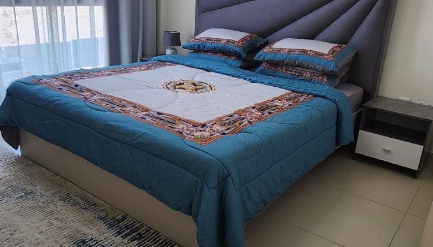 A bedroom with a double bed featuring a blue and patterned bedspread, two pillows, a nightstand, tiled floor, and a large window with gray curtains. A modern ceiling light is visible.