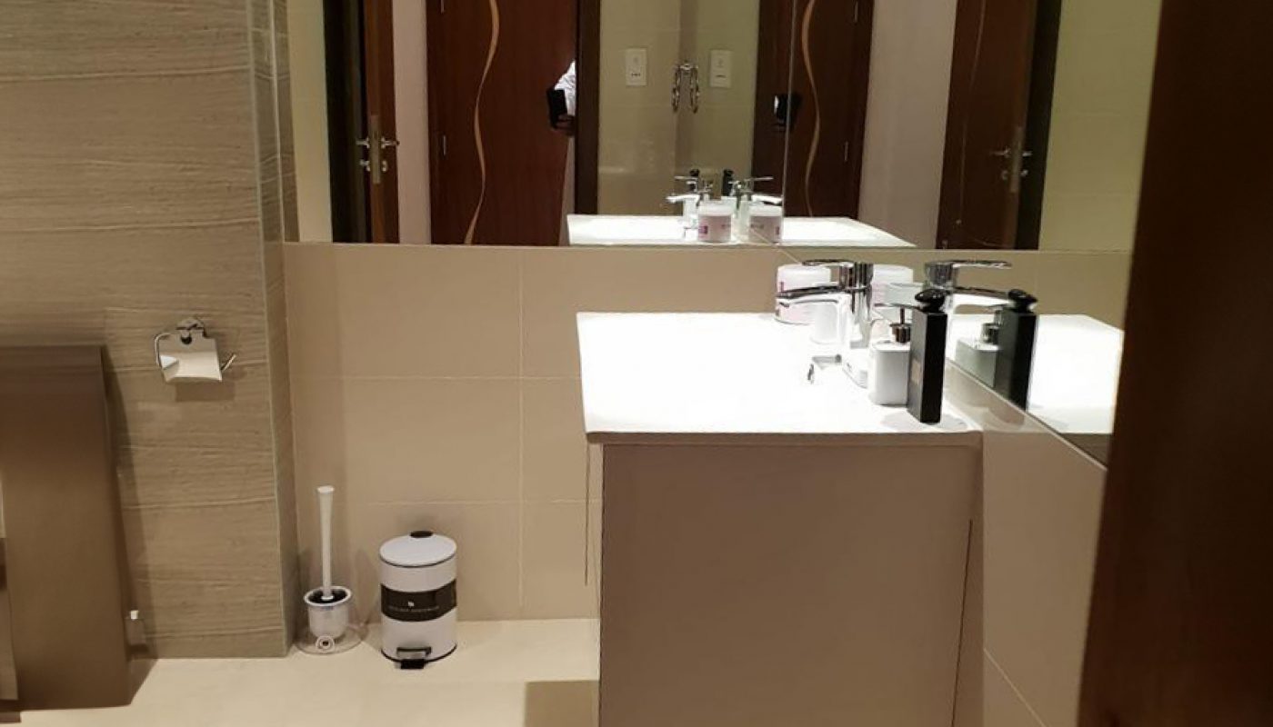 Modern bathroom with beige tiles, a wall-mounted sink, mirror, toilet, toilet paper holder, toilet brush, and a trash can. Two wooden doors and overhead lighting are visible.
