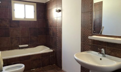 A bathroom with a bathtub, bidet, toilet, and sink. The walls and floor are tiled in dark brown, and there is a window above the bathtub. A mirror is mounted above the sink.