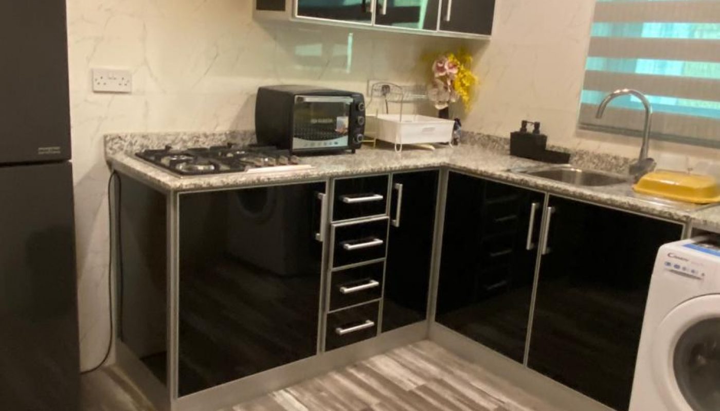 Modern kitchen with black cabinets, marble countertops, a fridge, microwave, gas stove, washing machine, and striped blinds. Pendant lights hang above the workspace.